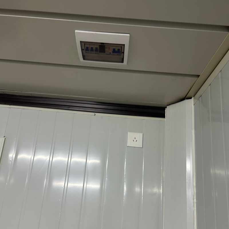 Interior detail of a folding container house showing a ceiling-mounted electrical circuit breaker box and a wall-integrated power outlet on white sandwich panels.
