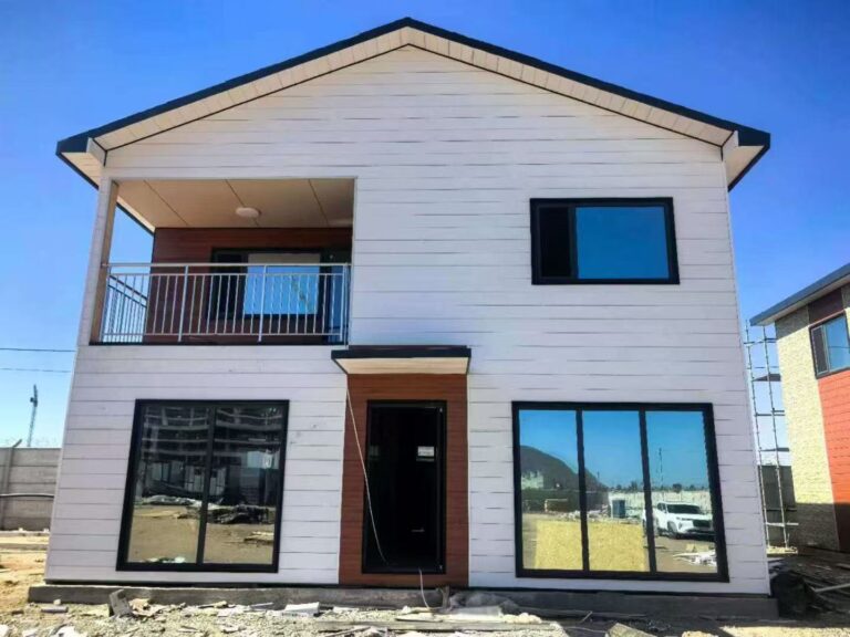 A white and wood-accented two-story detachable container house in Chile featuring large glass windows, a balcony with metal railings, and a sloped roof.
