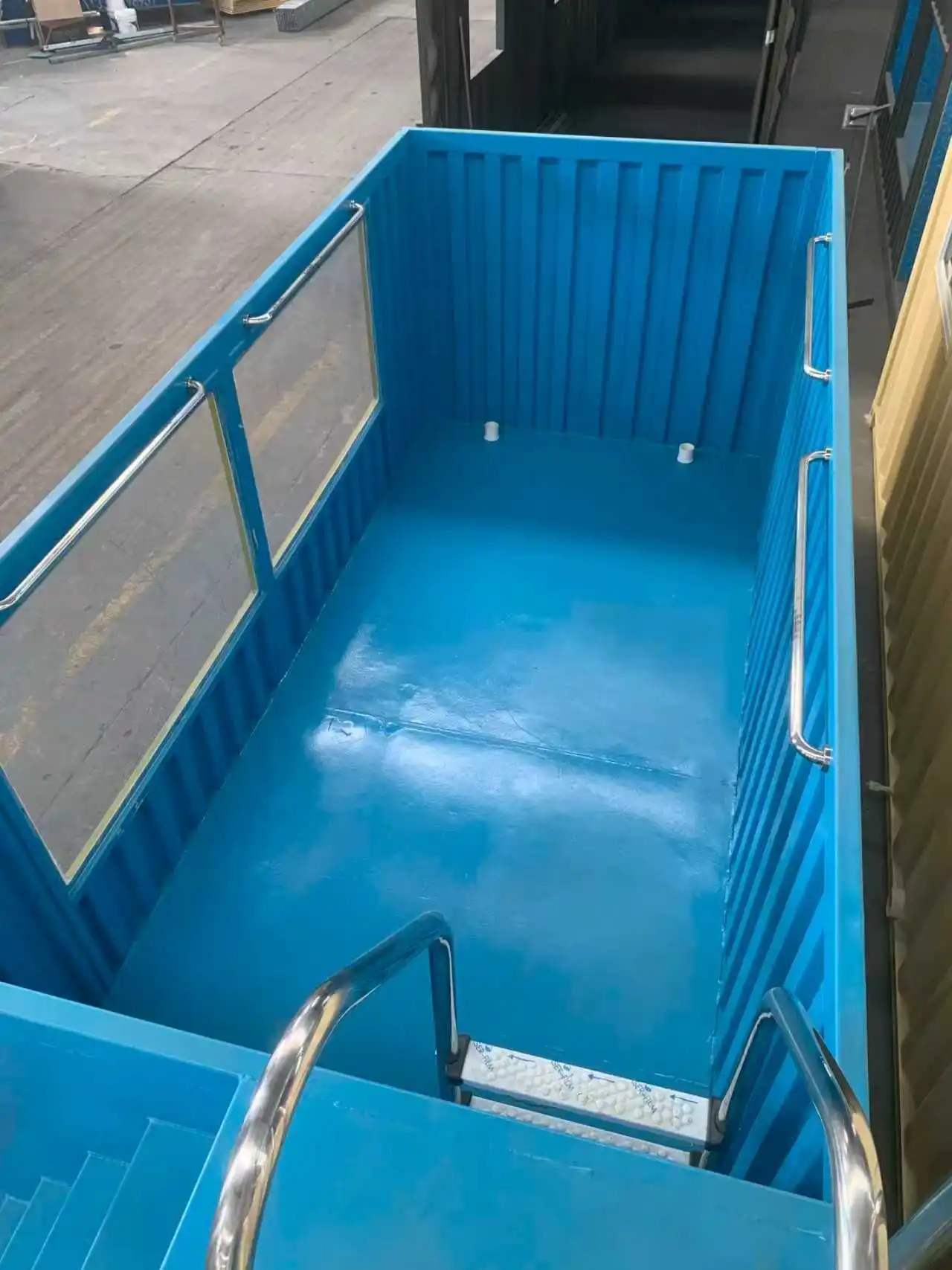 An interior view of a vibrant blue industrial shipping container swimming pool, featuring integrated clear glass viewing windows, stainless steel safety handrails, and built-in steps for easy access.