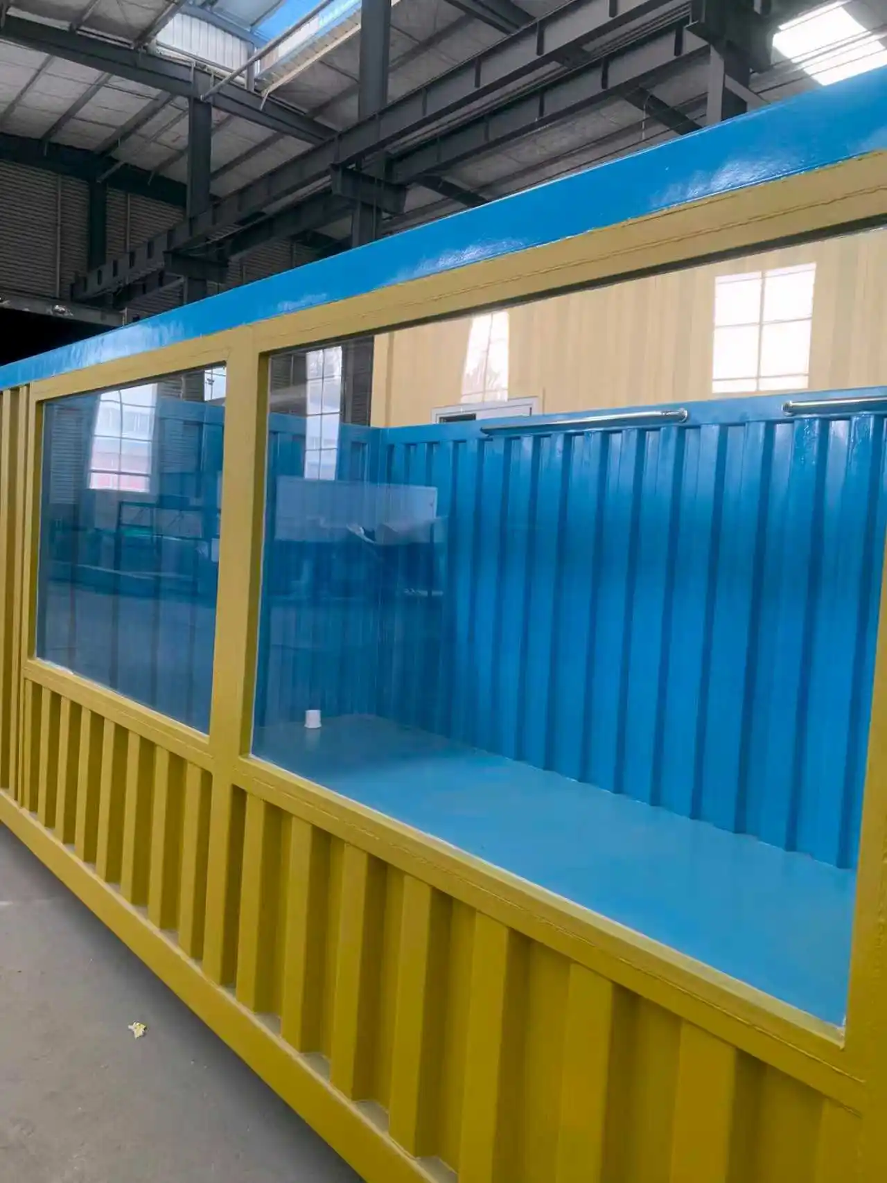 A side-angle view of a premium container swimming pool inside a factory, showcasing its yellow corrugated steel exterior and a large, high-strength transparent glass viewing window that reveals the vibrant blue waterproof interior.