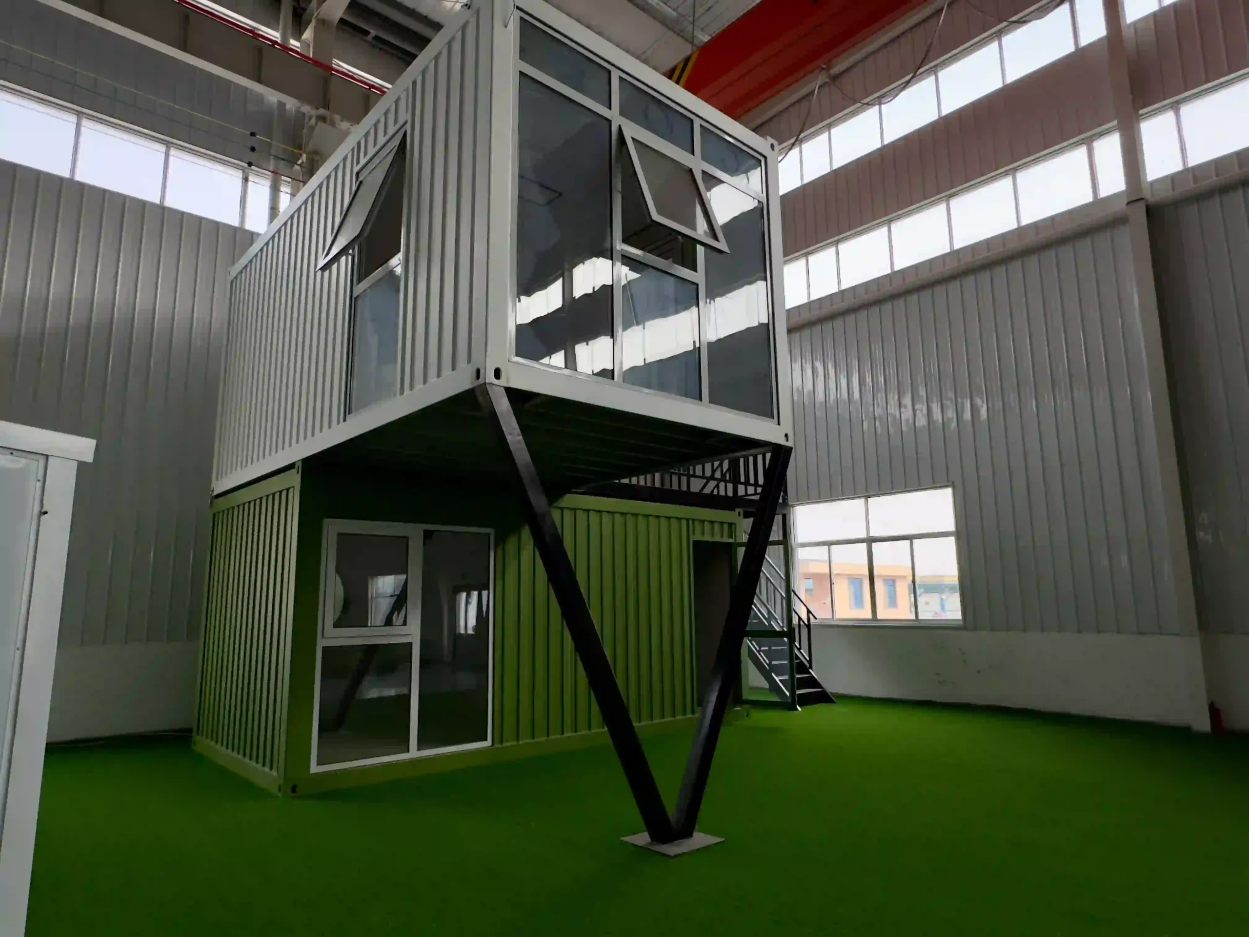 A modern two-story stacked shipping container house featuring a cantilevered upper level, integrated floor-to-ceiling glass windows, a private second-floor balcony with safety railings, and a sleek dark-grey industrial finish.